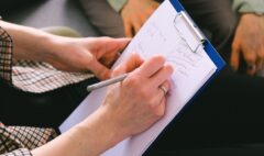 Close-up of a professional writing client notes on a clipboard during a conversation.