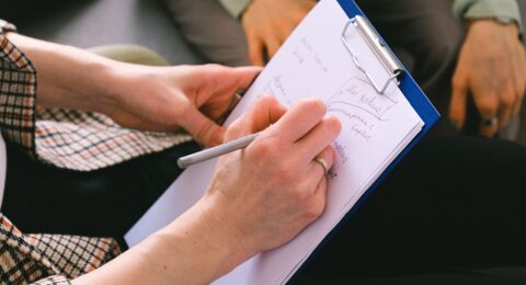 Close-up of a professional writing client notes on a clipboard during a conversation.