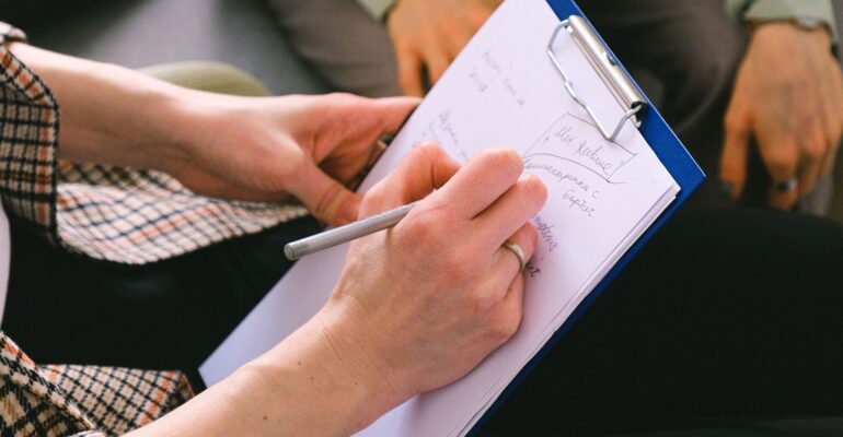 Close-up of a professional writing client notes on a clipboard during a conversation.