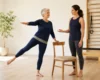 An active older adult practising a supported standing balance task while a Pilates instructor observes nearby