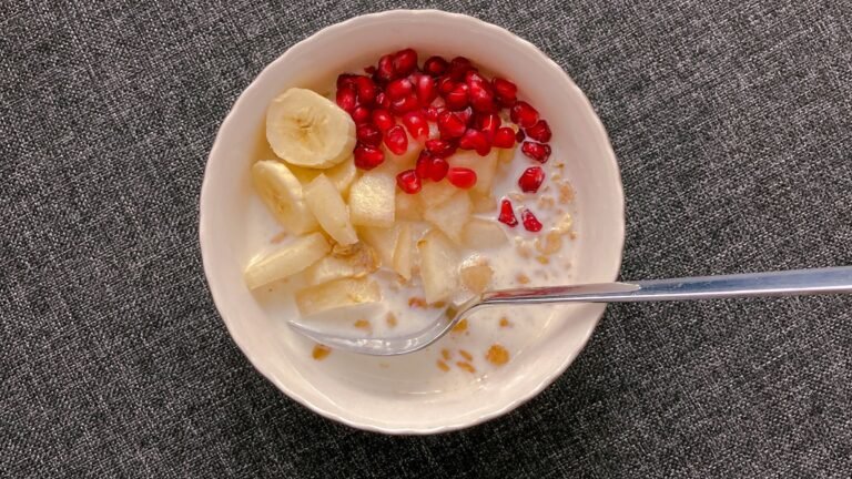 A bowl of oats with milk, banana, apple and pomegranate, viewed from above.