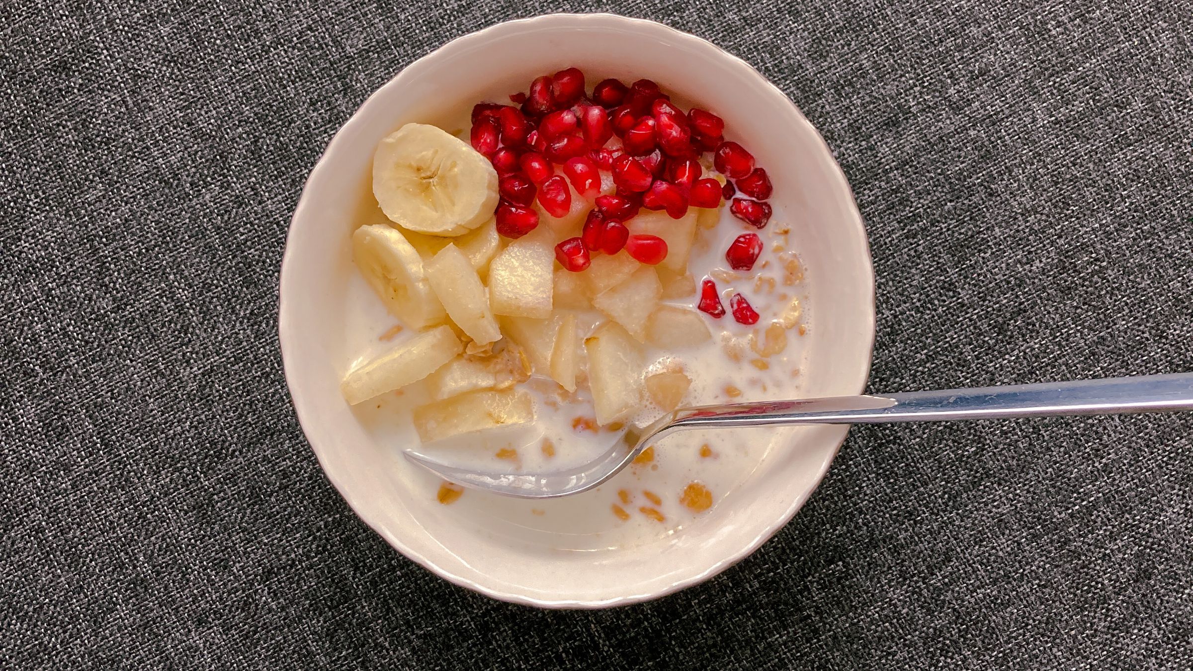 A bowl of oats with milk, banana, apple and pomegranate, viewed from above.