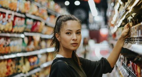 An adult shopper considering products in a supermarket aisle filled with packaged foods.