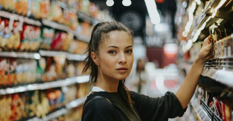 An adult shopper considering products in a supermarket aisle filled with packaged foods.