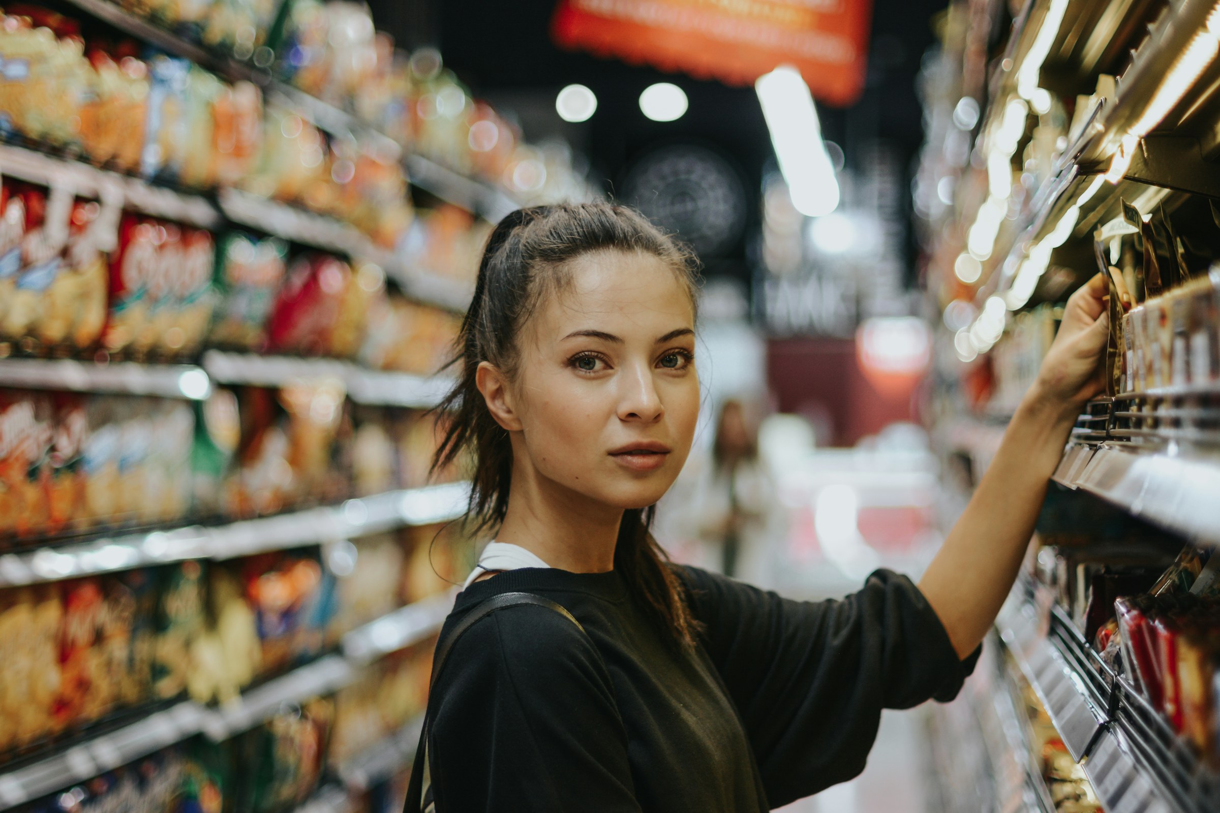 An adult shopper considering products in a supermarket aisle filled with packaged foods.