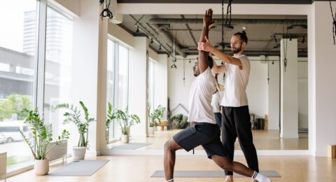 Yoga teacher guiding an adult student through a controlled standing pose in a bright studio