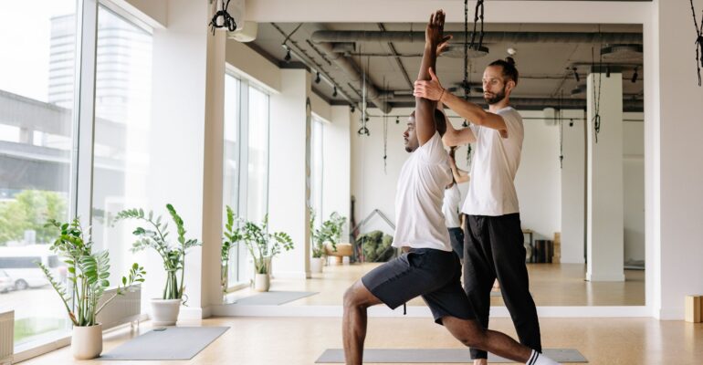Yoga teacher guiding an adult student through a controlled standing pose in a bright studio