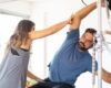 An instructor guides a man's side bend beside Pilates apparatus in a bright studio.