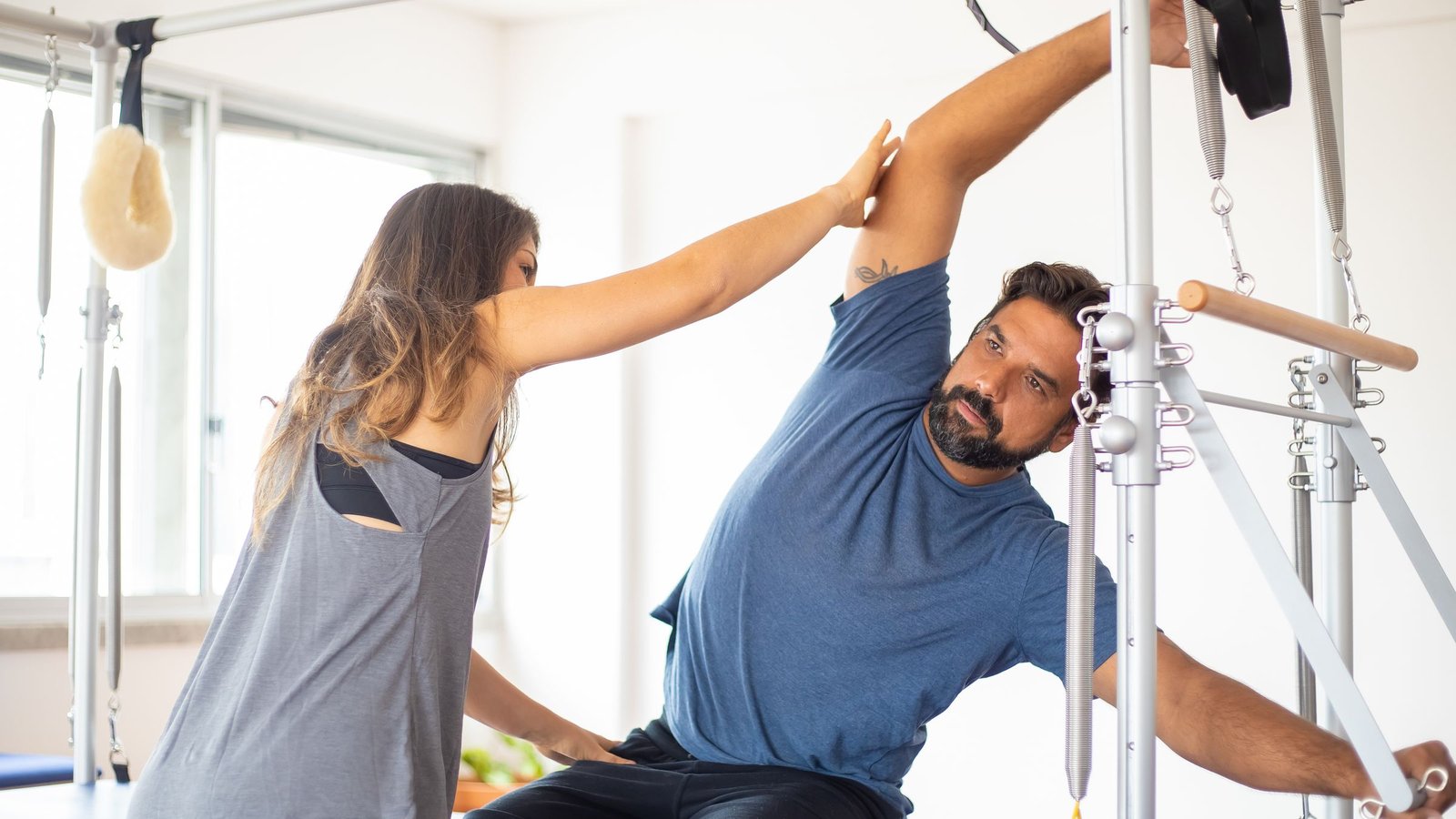 An instructor guides a man's side bend beside Pilates apparatus in a bright studio.