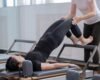 An instructor gives hands-on guidance to an adult learner during a reformer Pilates movement.